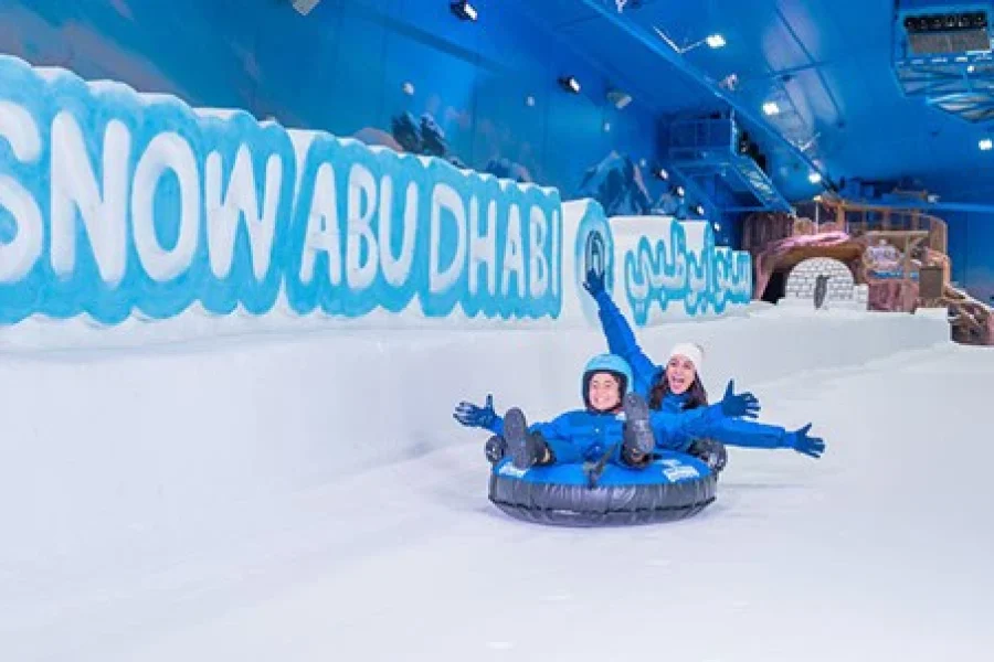 A woman and child laughing while sliding down a snow-covered slope on a black inner tube at Snow Abu Dhabi, with a large ice-carved sign of the park's name in the background.