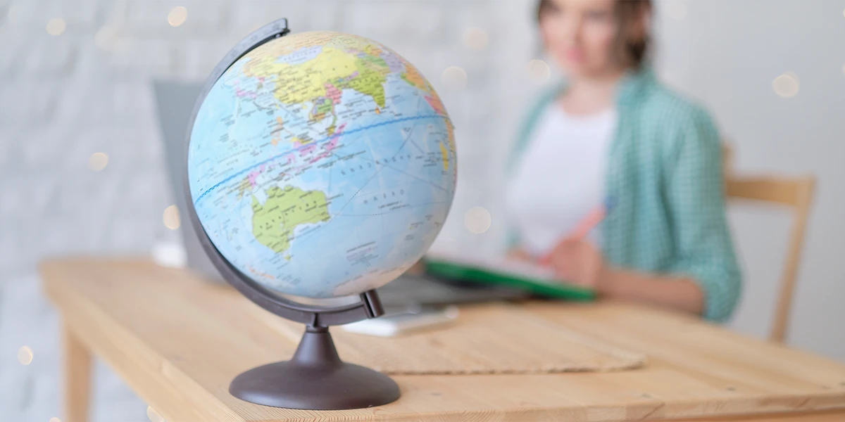 A world globe stands in focus on a wooden table, while a person in the background plans a trip, illustrating the choice between International Tour Companies vs Solo Travel: What Works Better for global explorers.