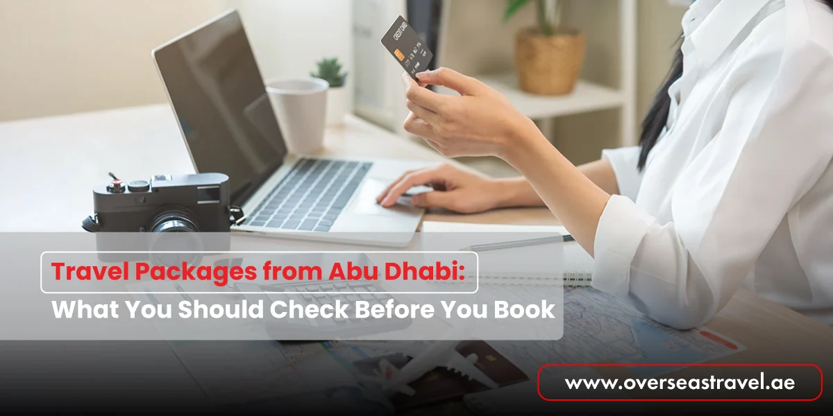A woman using a credit card and laptop to book travel packages from Abu Dhabi, with a camera and maps on the desk suggesting holiday planning.