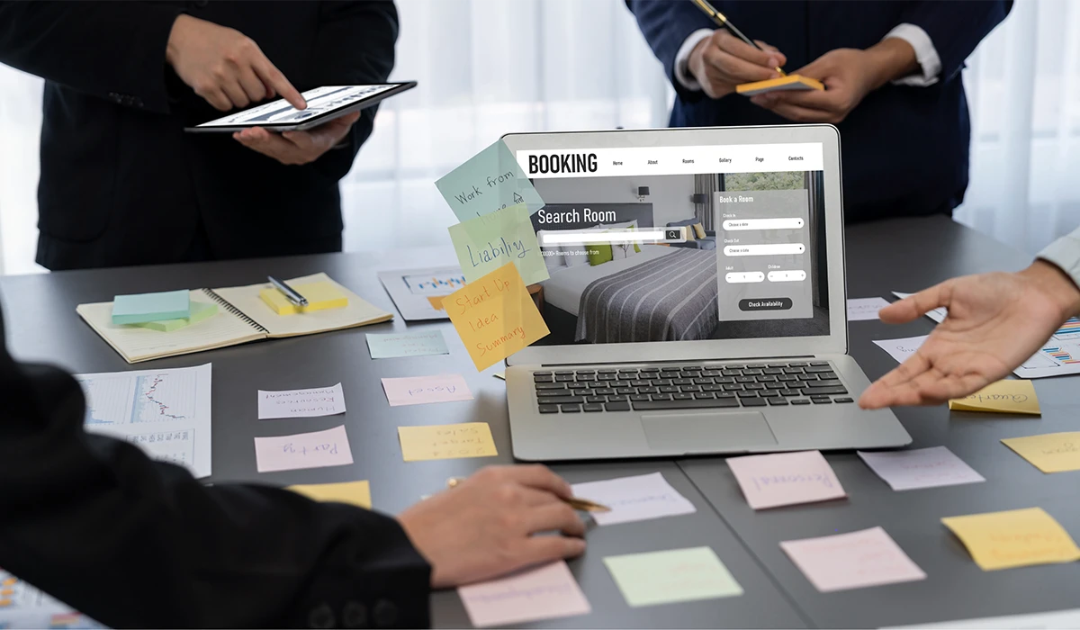 Corporate Travel Booking planning meeting with business professionals reviewing an online hotel reservation system on a laptop, surrounded by notes and documents.